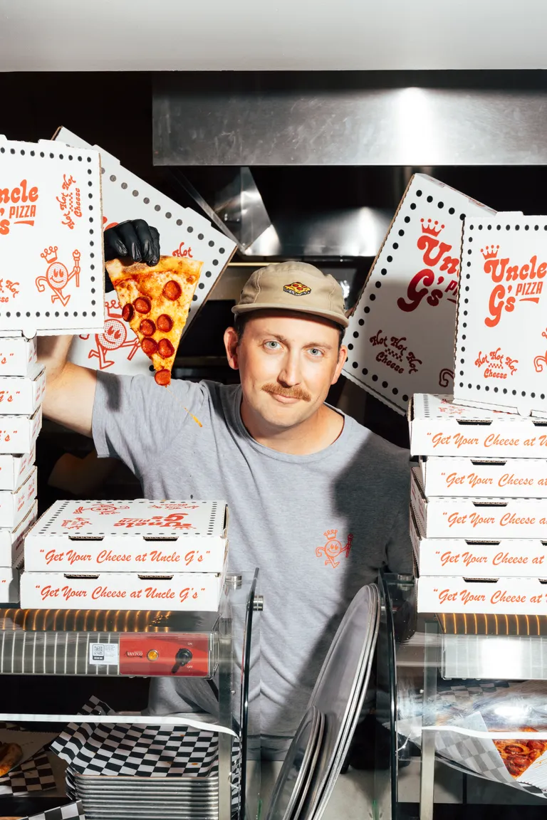 George holding up a pepperoni slice between stacks of Uncle G's pizza boxes