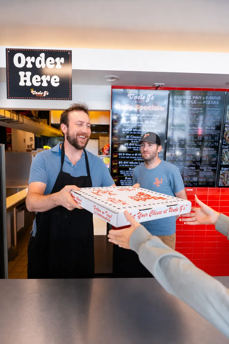 George handing a pizza box to a customer under an Order Here sign