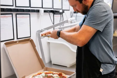 George boxing a finished pizza at the prep bench beneath the kitchen task charts