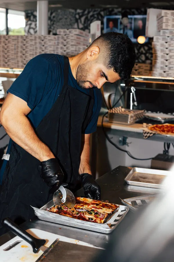 A line cook slicing a Detroit-style pizza with a cutting wheel at the pass