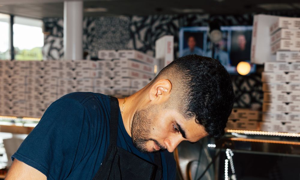 An Uncle G's line cook in a black apron looking down at his work at the pass, stacked pizza boxes behind him