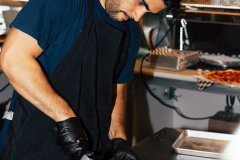 An Uncle G's line cook in a black apron looking down at his work at the pass, stacked pizza boxes behind him