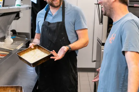 George mid-conversation with a laughing teammate in a trucker cap in the shop kitchen